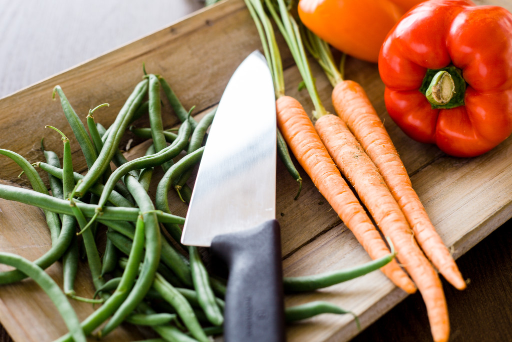 Side angle shot of chef's knife illuminated on wooden platter with fresh green beans, raw carrots, and red and orange bell pepper.