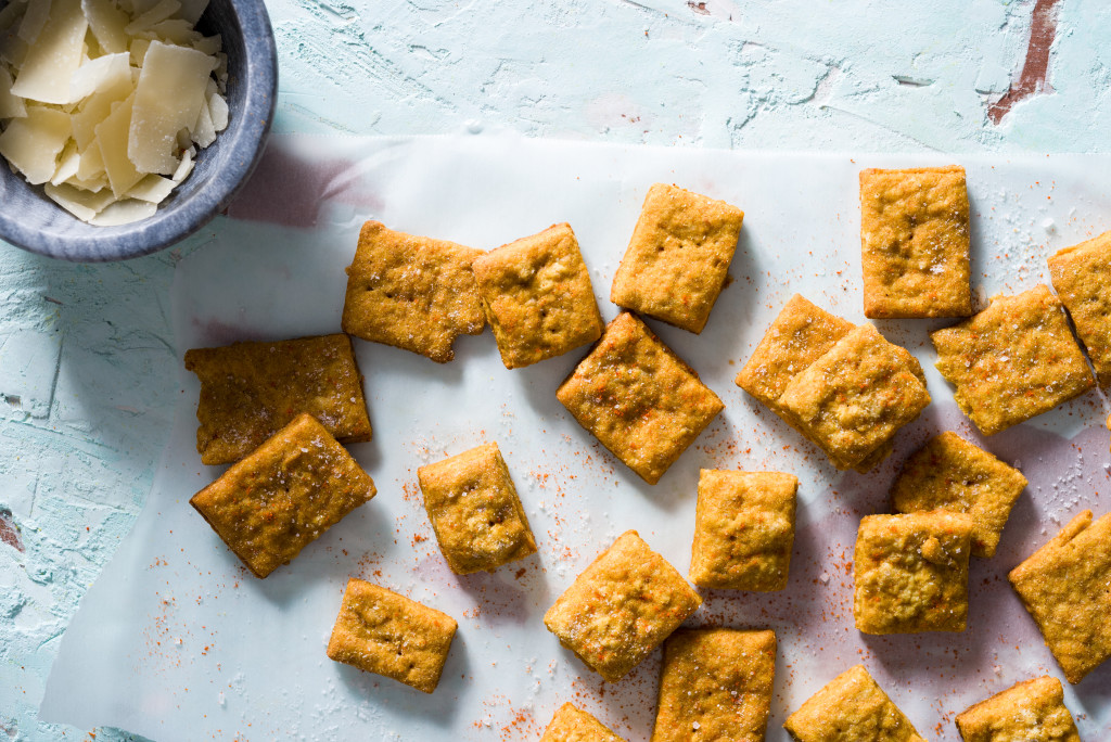 Close-up of homemade cheesy turmeric crackers on a textured blue paster surface with copper accents and small bowl of shaved parmesan cheese.