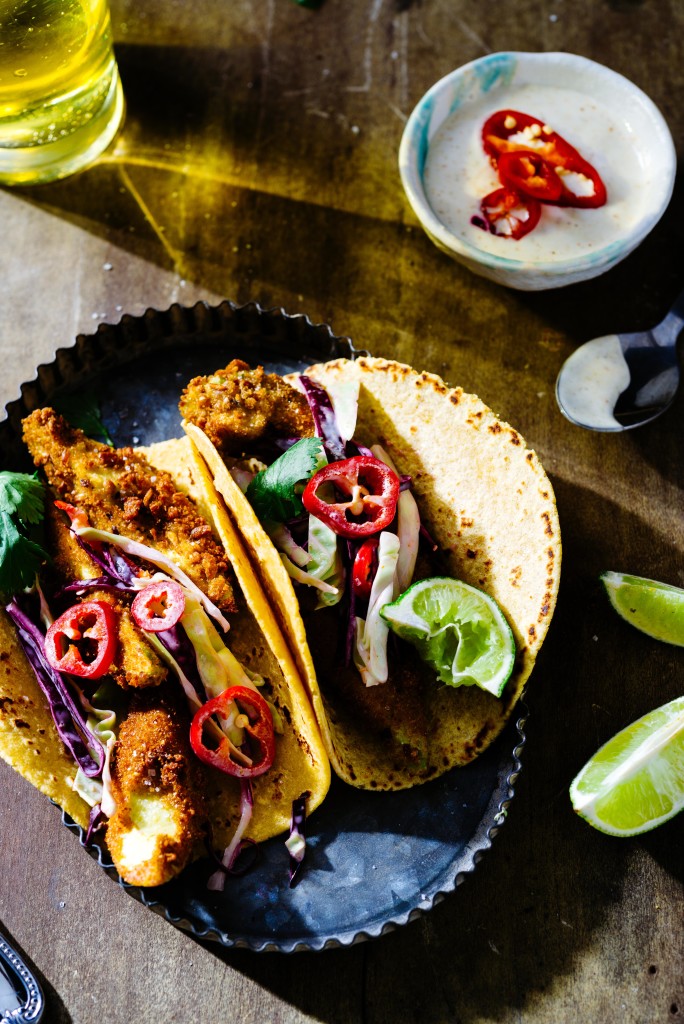 Overhead of fried avocado tacos with red cabbage slaw and sliced red jalapenos in metal tray with lime wedges, creamy sauce and beer on wooden surface.