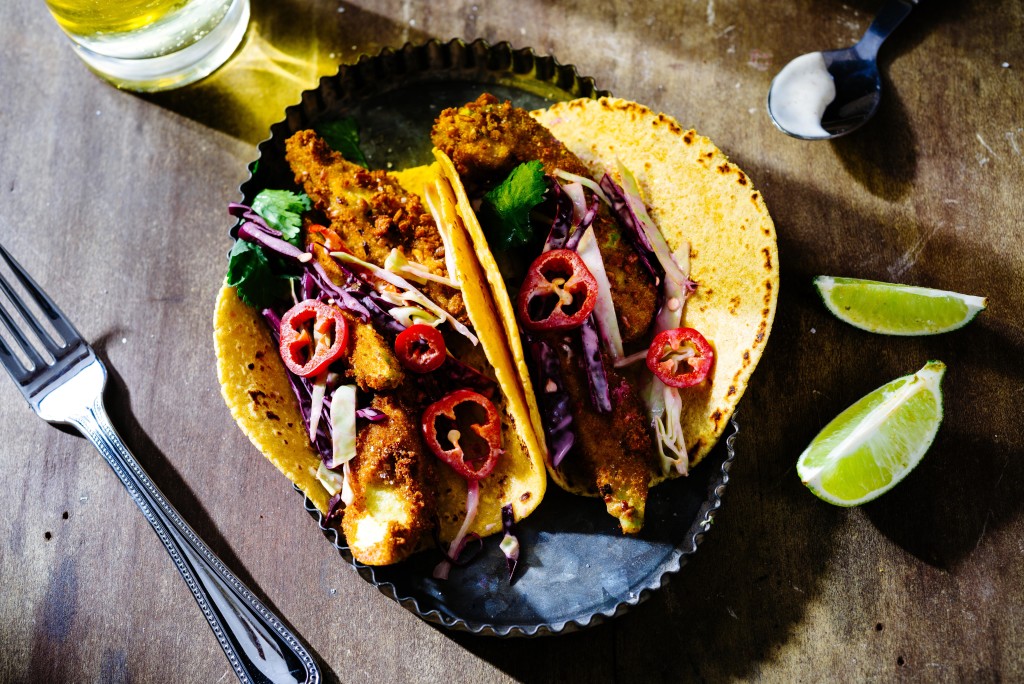 Overhead of fried avocado tacos with red cabbage slaw and sliced red jalapenos in metal tray with lime wedges and beer on wooden surface.