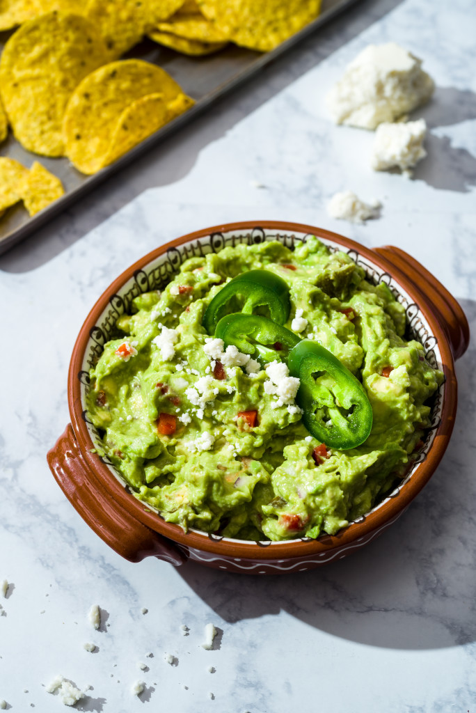 Brown ceramic bowl of chunky guacamole with pimentos, queso fresco and sliced jalapenos on white marble surface with metal tray of tortilla chips in the background.