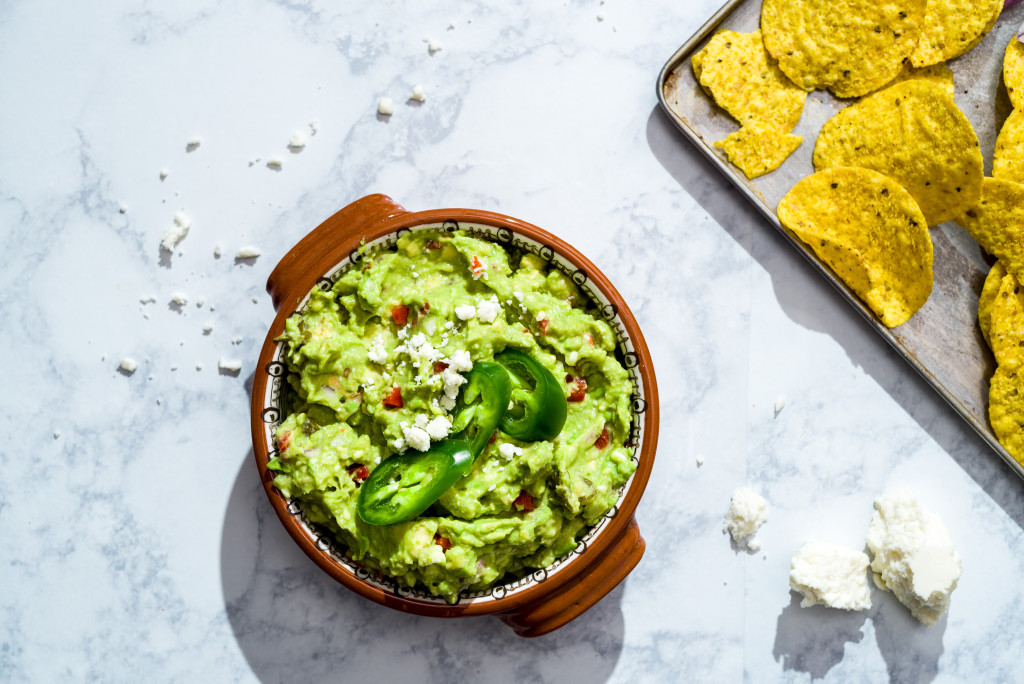 Overhead of brown ceramic bowl of chunky guacamole with pimentos, queso fresco and sliced jalapenos on white marble surface with metal tray of tortilla chips in the background.